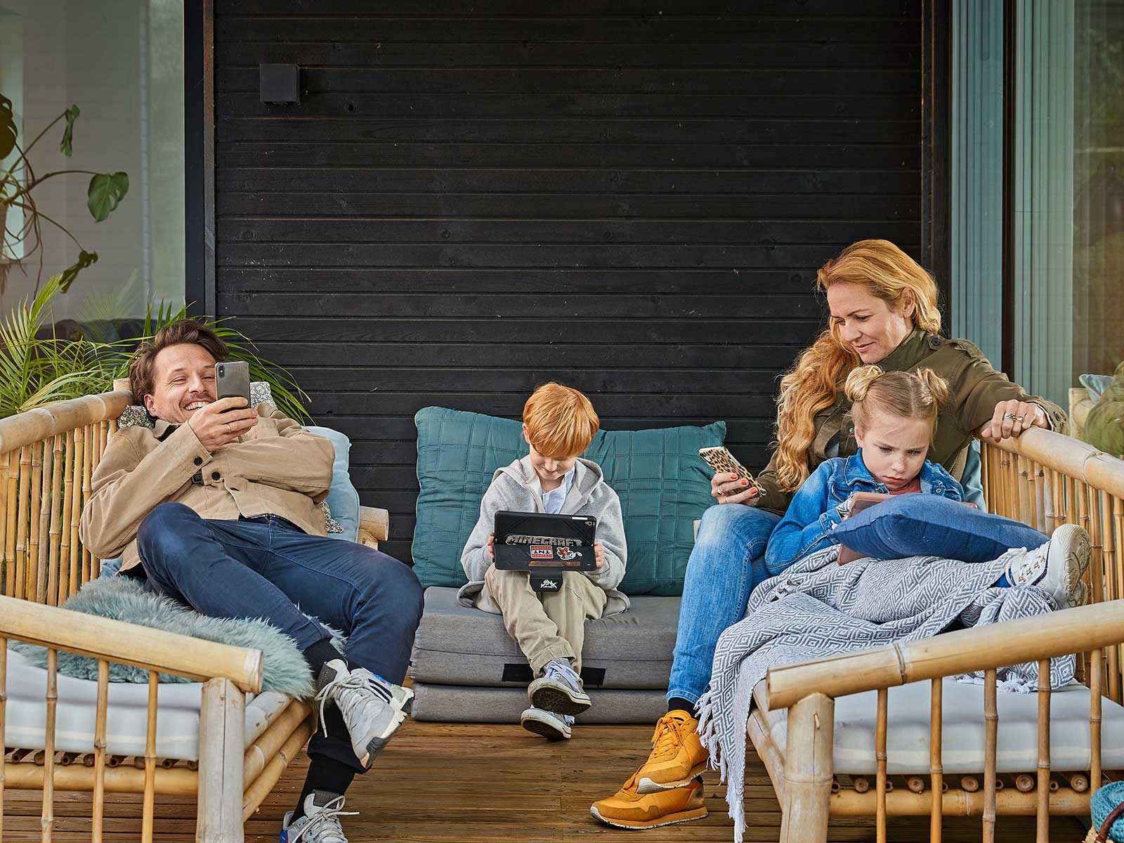 Familie på fire sidder på terrassen og spiller på ipad og mobiler. De er meget tilfredse med deres fibernet priser fra EWII.